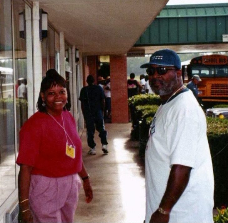 Two smiling alumni outside a building at the 1999 reunion, with a school bus in the background.
